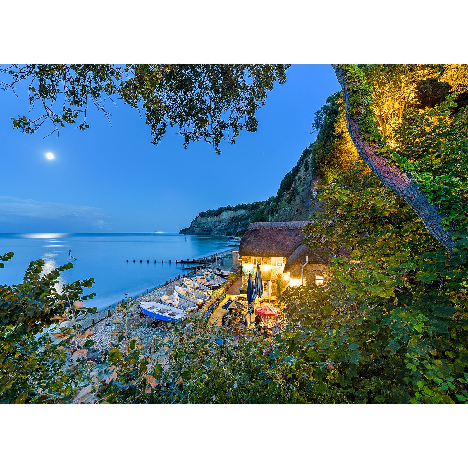 A tranquil evening with a Shanklin by Moonlight over a coastal cove on the Isle of Wight, featuring boats and a thatched-roof structure captured by Available Light Photography.