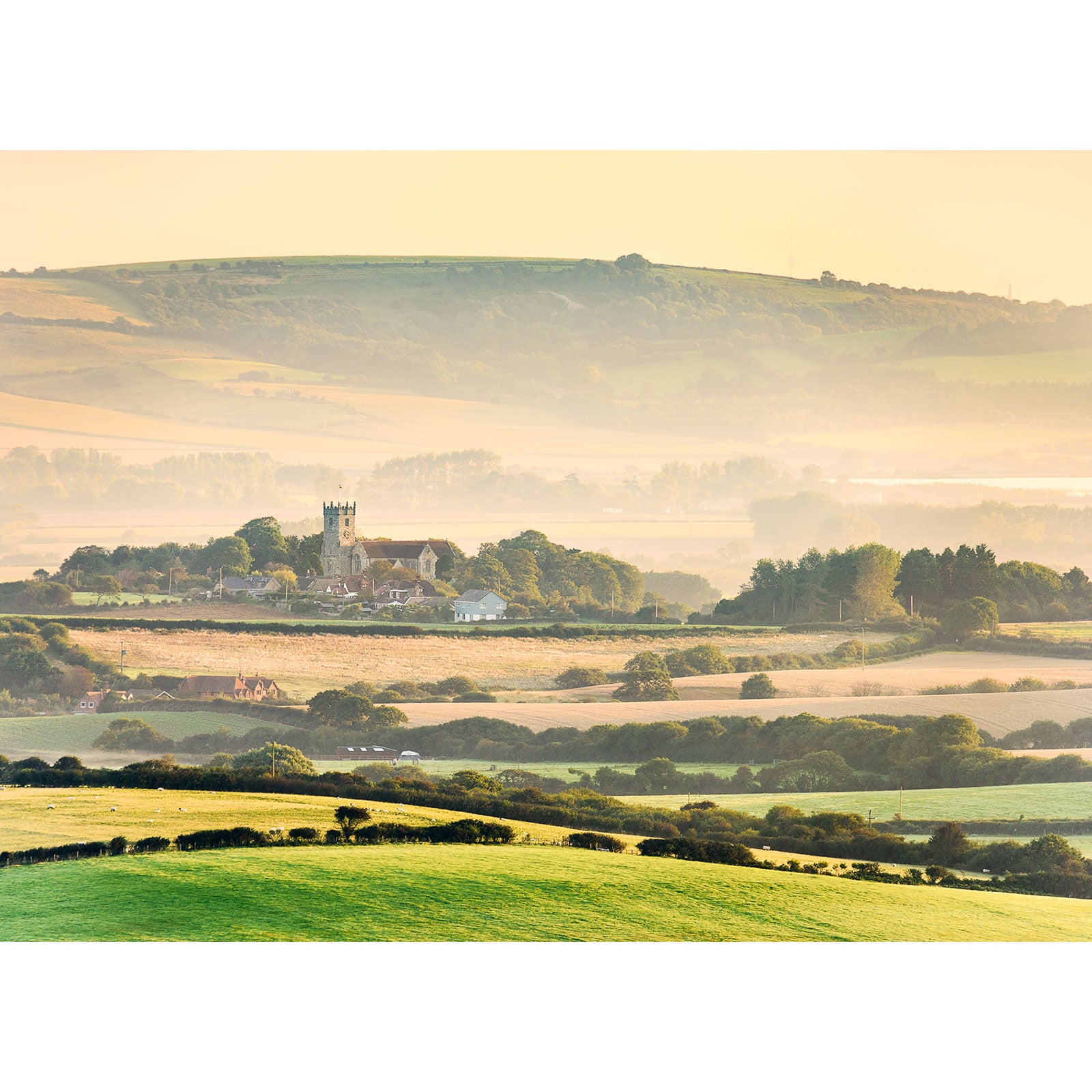 Rolling hills with a Godshill Church amidst a rural landscape on the Isle of Wight, bathed in soft, hazy sunlight captured by Available Light Photography.