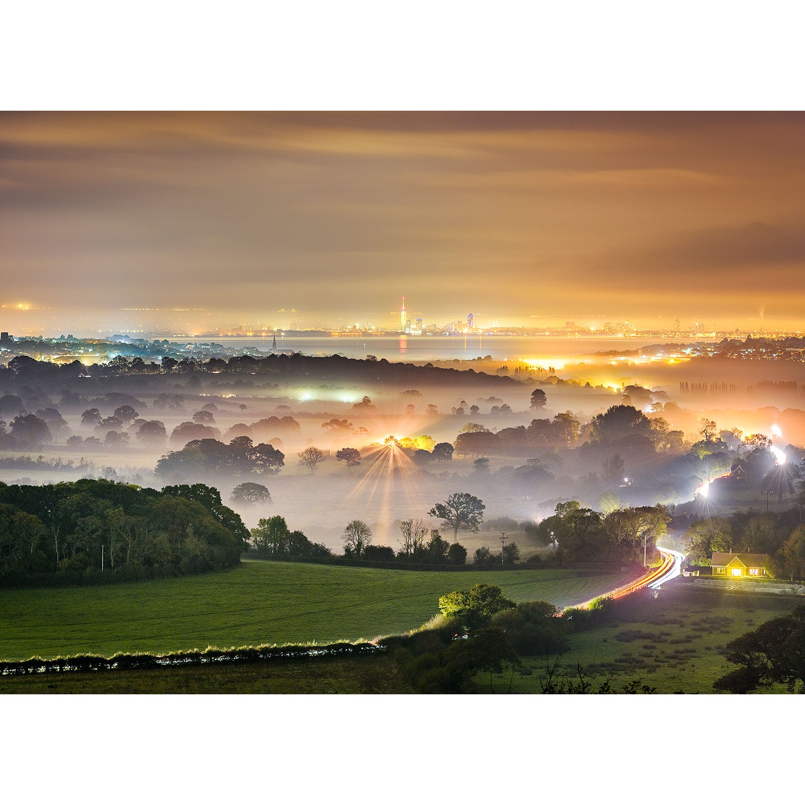 A nocturnal landscape blanketed in Misty Evening with twinkling city lights on the Isle of Wight in the distance and a winding road illuminated by a vehicle's passing, captured by Available Light Photography.