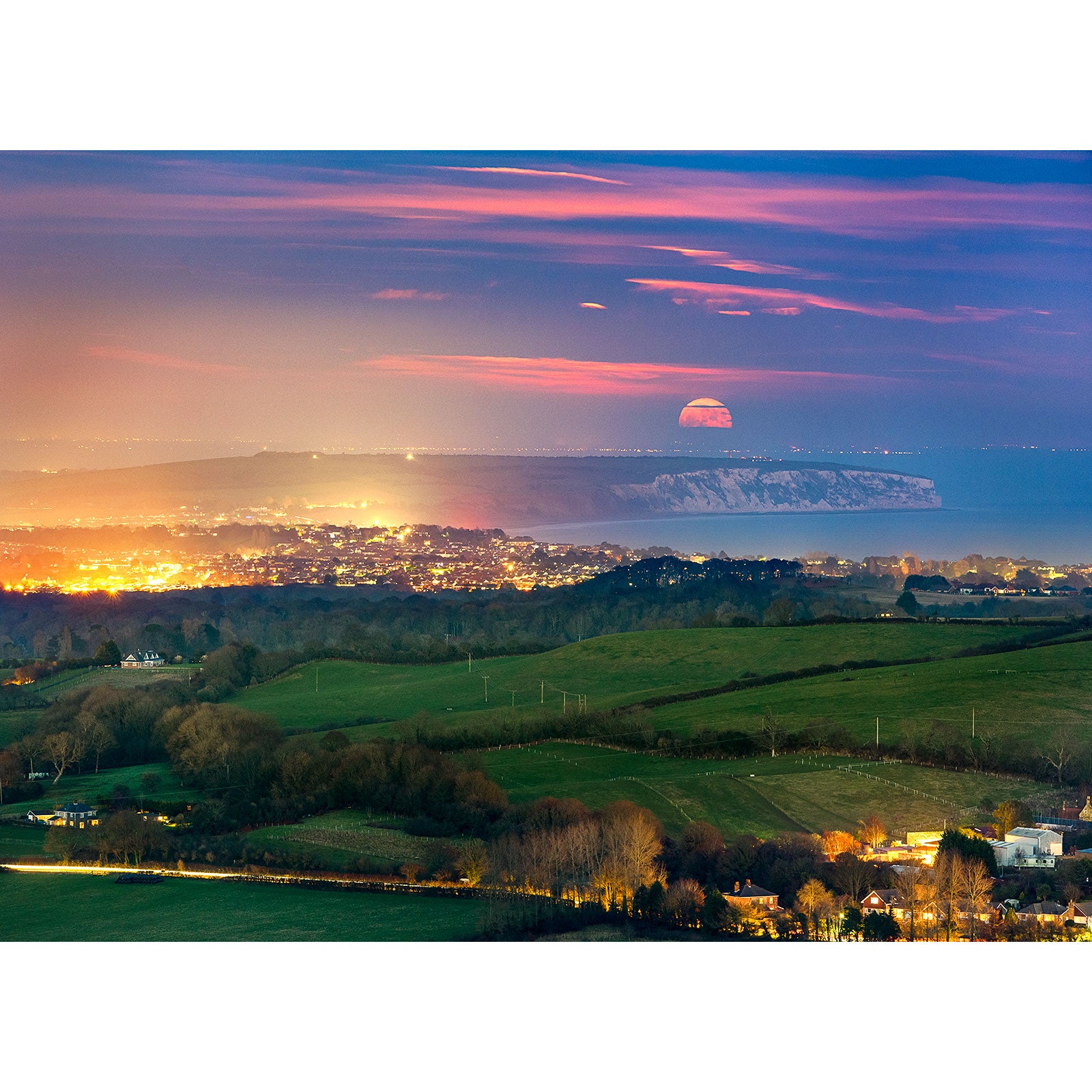 A Moonrise over Sandown Bay rising over a coastal landscape at twilight, with city lights twinkling below on the Isle of Wight. (Available Light Photography)