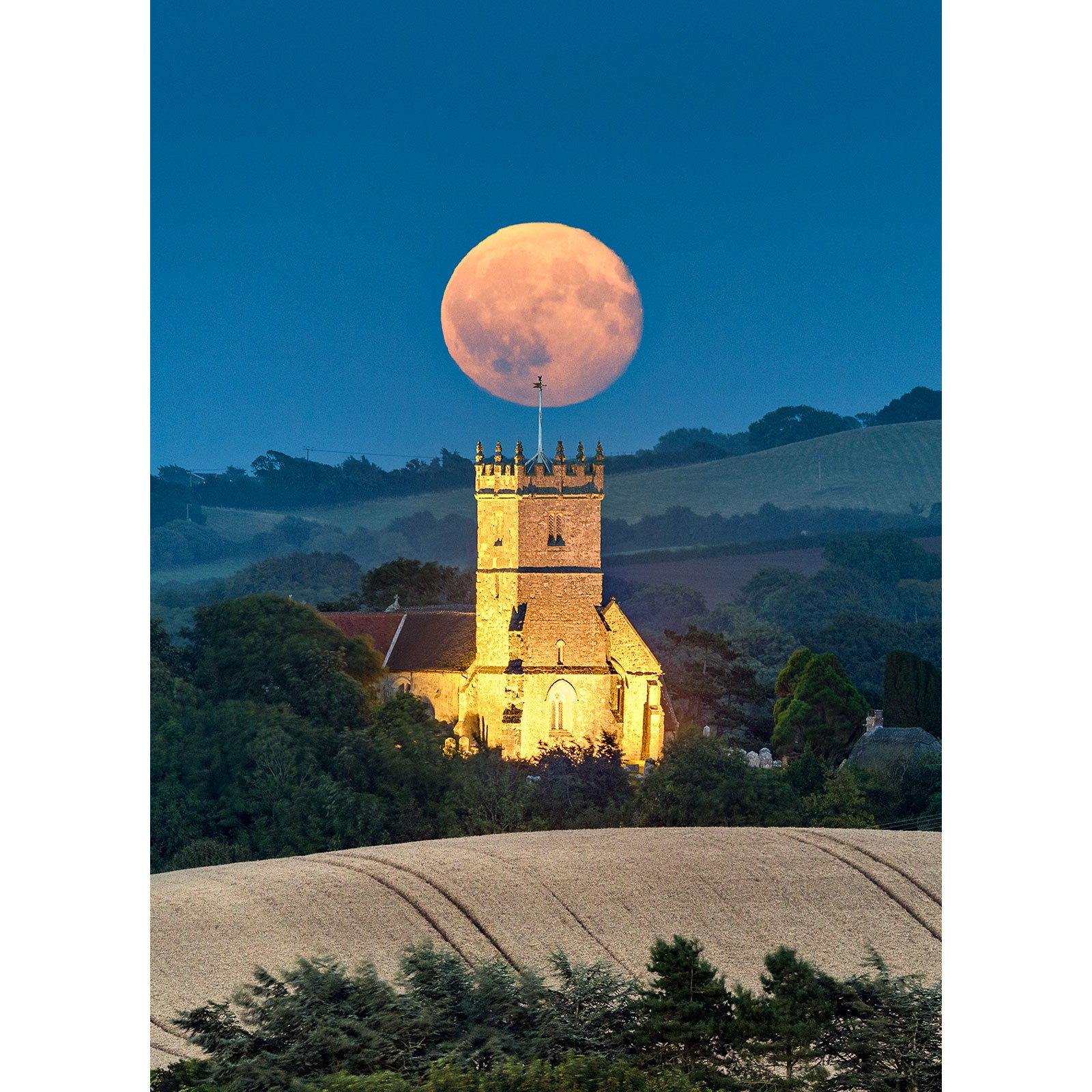 A Moonrise, Godshill Church rising behind an illuminated church set against hills in the twilight on the Isle of Gascoigne by Available Light Photography.