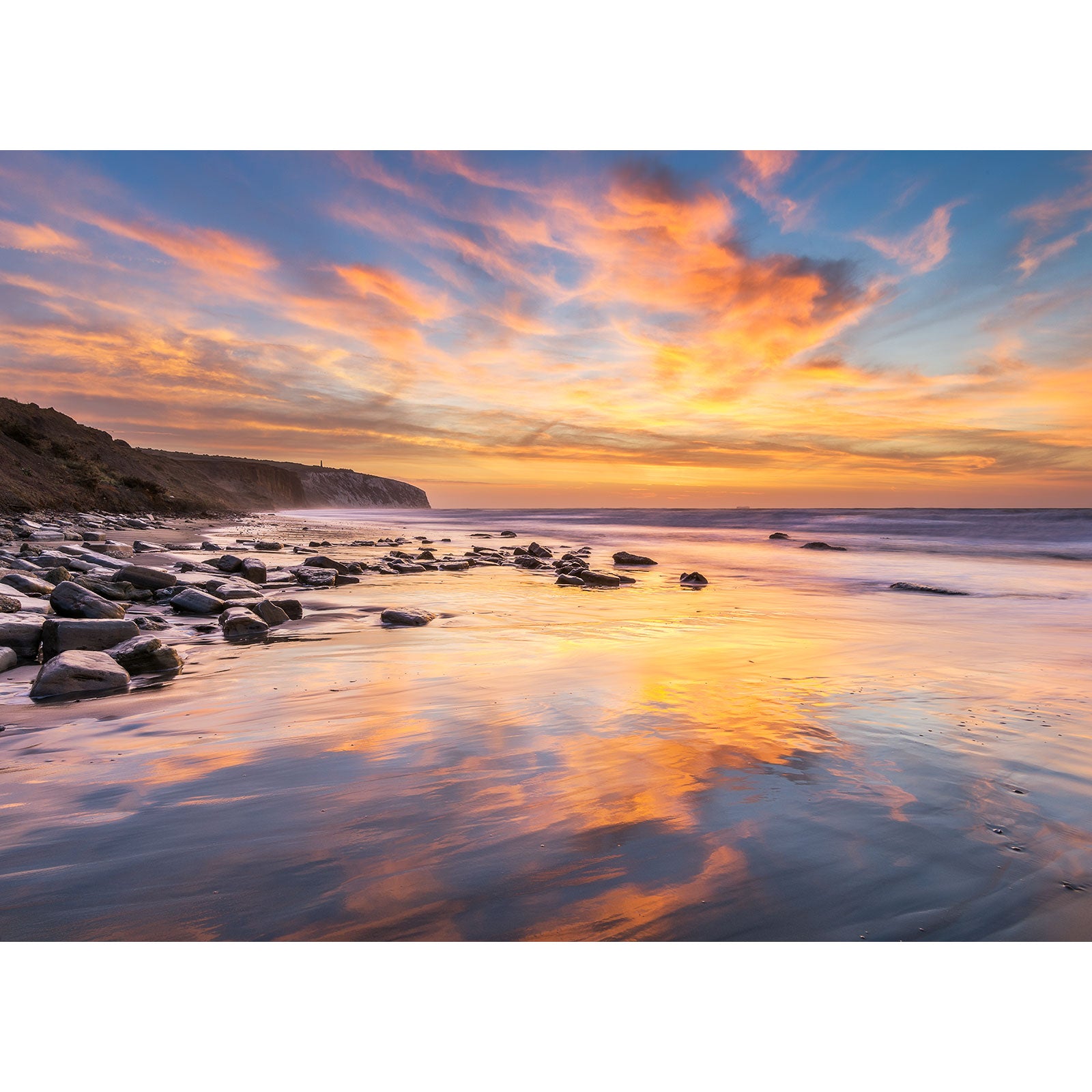 Sunset over Culver Cliff, a calm beach on Wight with reflections on the water and scattered rocks on the sand, captured by Available Light Photography.