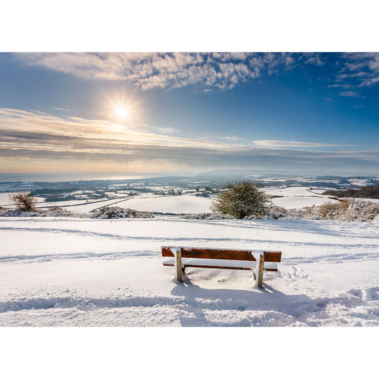 Serene winter landscape with a bench overlooking Snow over Sandown Bay-covered hills under a clear sky with a bright sun, painted by Steve. (Brand: Available Light Photography)