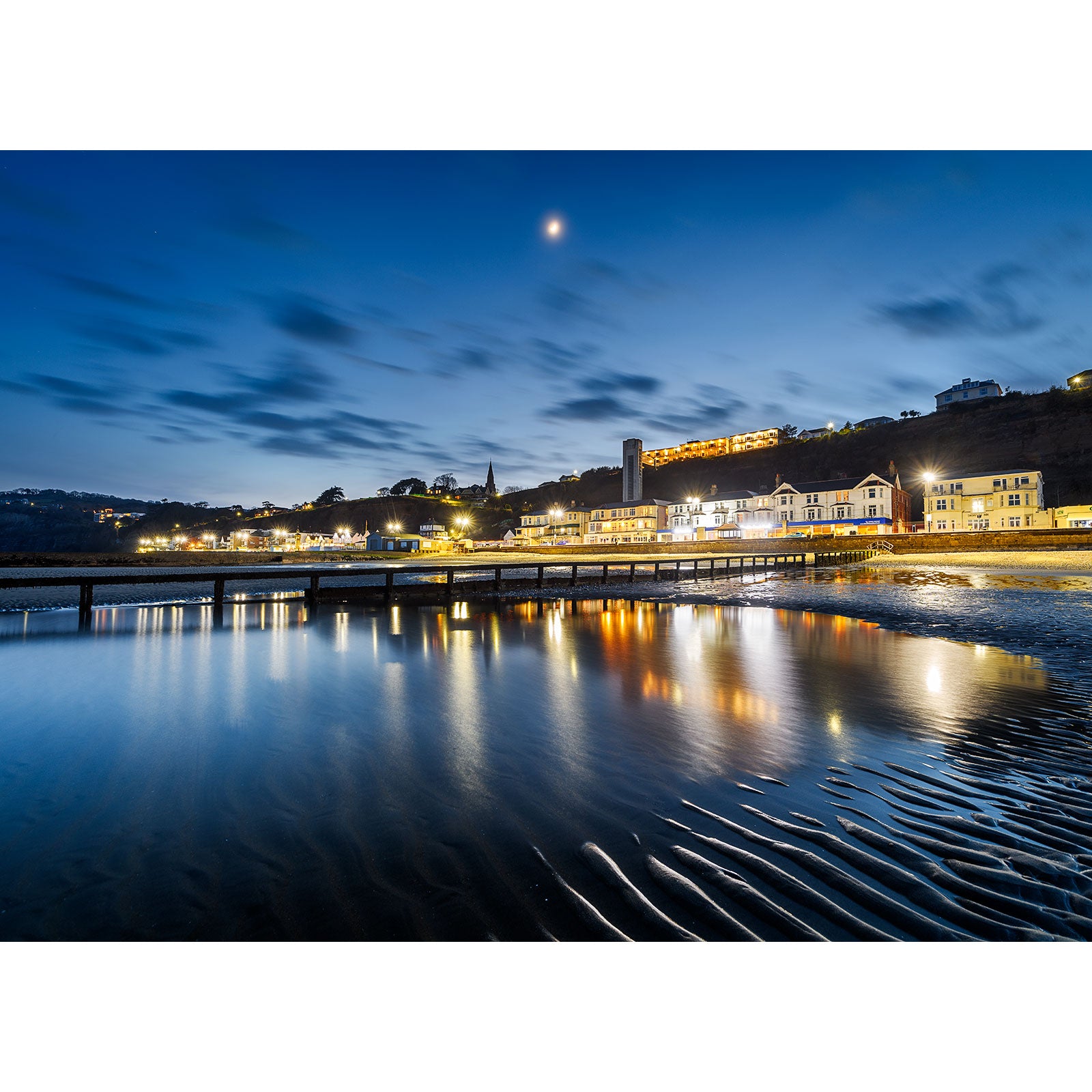 Twilight over Shanklin Beach on the Isle of Wight with reflections on the water, sand patterns in the foreground, and a moon above, photographed by Available Light Photography.