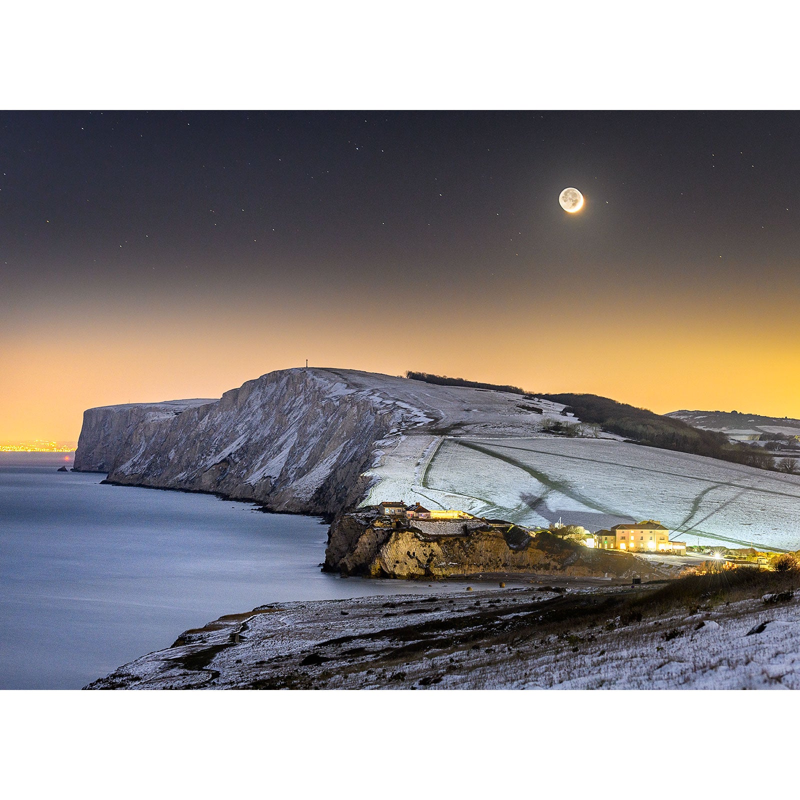 A Crescent Moon, Tennyson Down coastal landscape with snow-covered cliffs and a small illuminated village on the Isle at night by Available Light Photography.