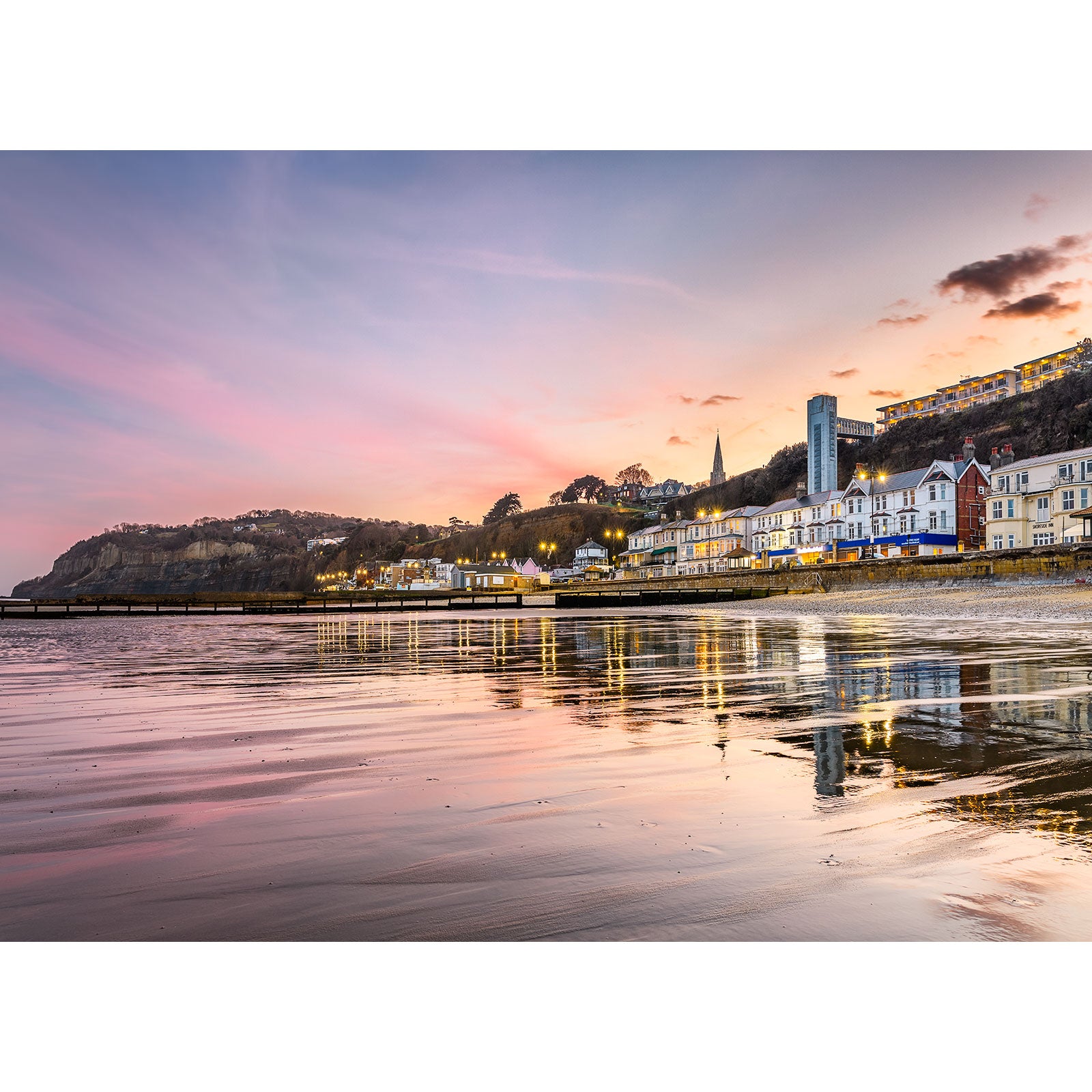Coastal town at sunset with buildings reflecting on Shanklin Beach water by Available Light Photography.