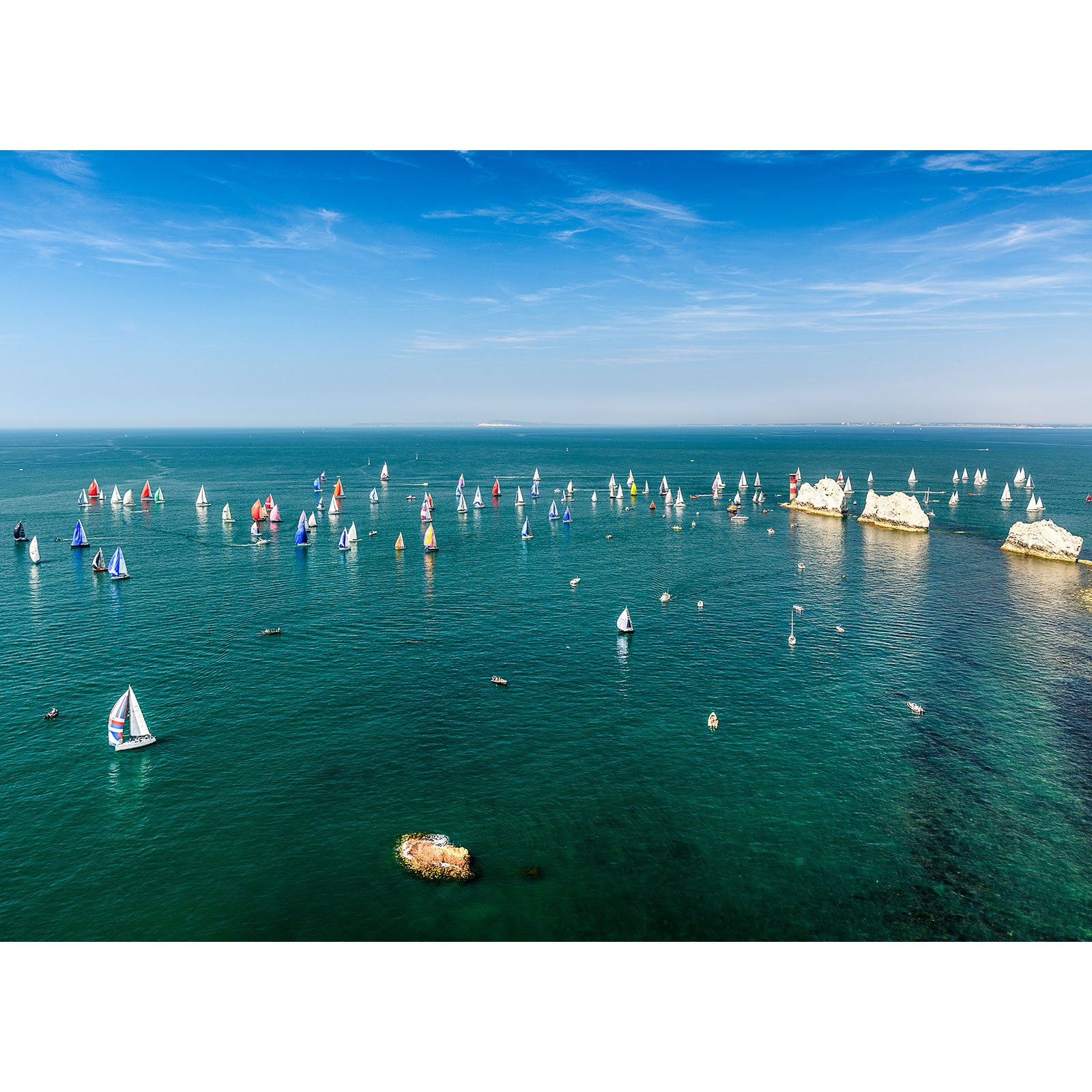 Aerial view of the Round the Island Race near the Isle of Gascoigne with numerous sailboats spread out over open water, with a clear blue sky above and rocky outcrops visible in the sea by Available Light Photography.