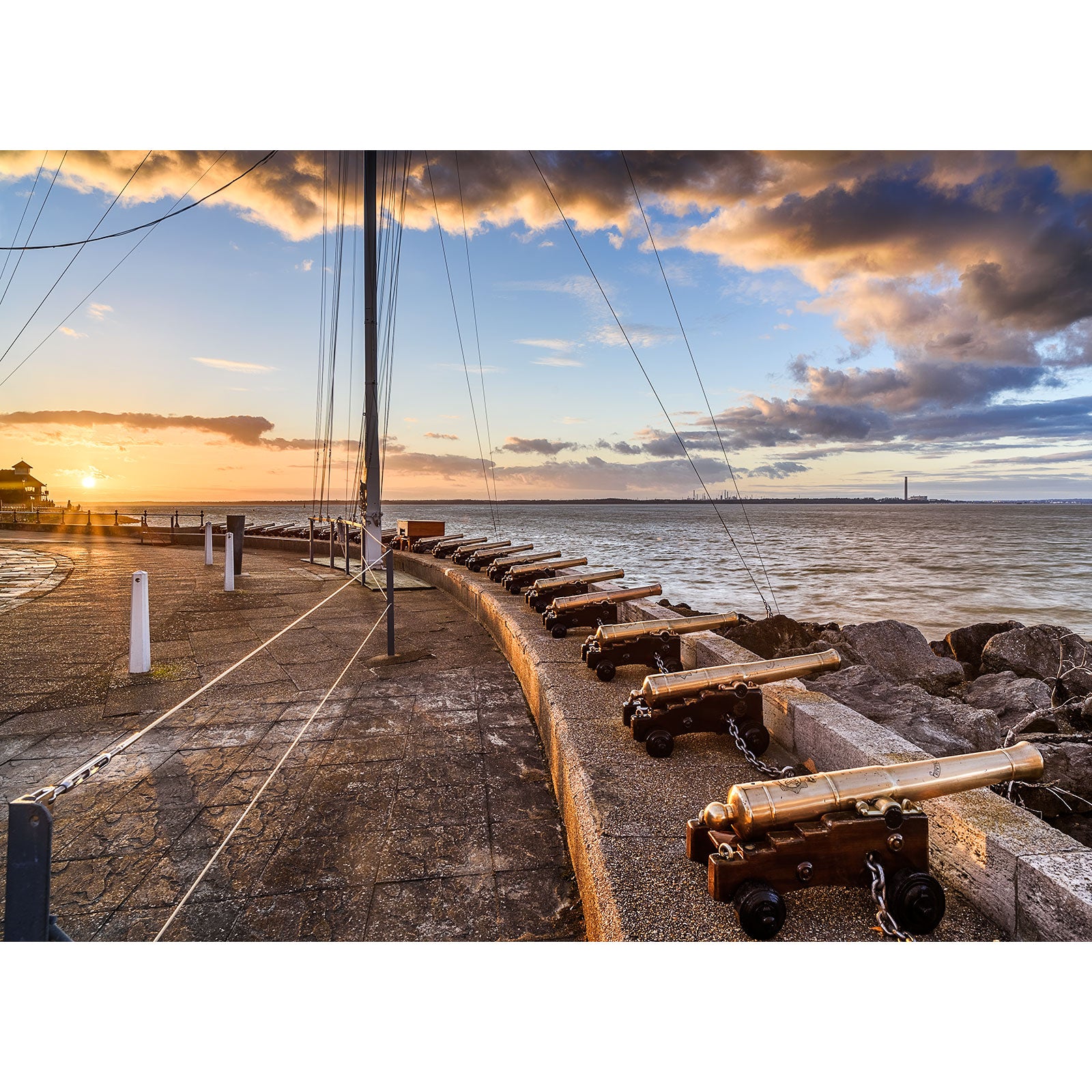 Sunset at a waterfront with a row of Cowes cannons, overlooking a calm sea with the Isle of Wight bridge in the distance, captured by Available Light Photography.