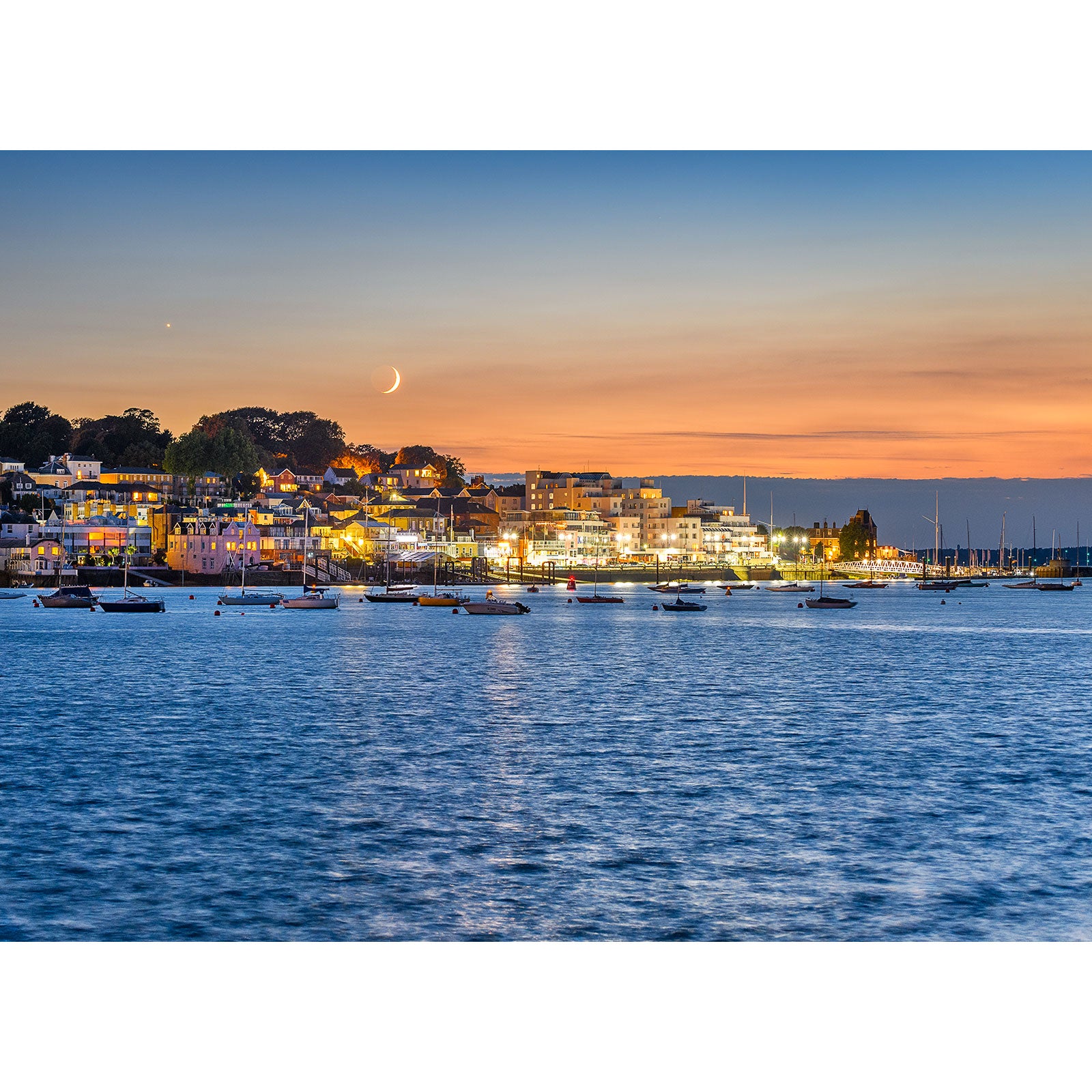 Twilight over a coastal town on the Isle of Wight with "Crescent Moon over Cowes" above, lights reflecting on water. Created by Available Light Photography.