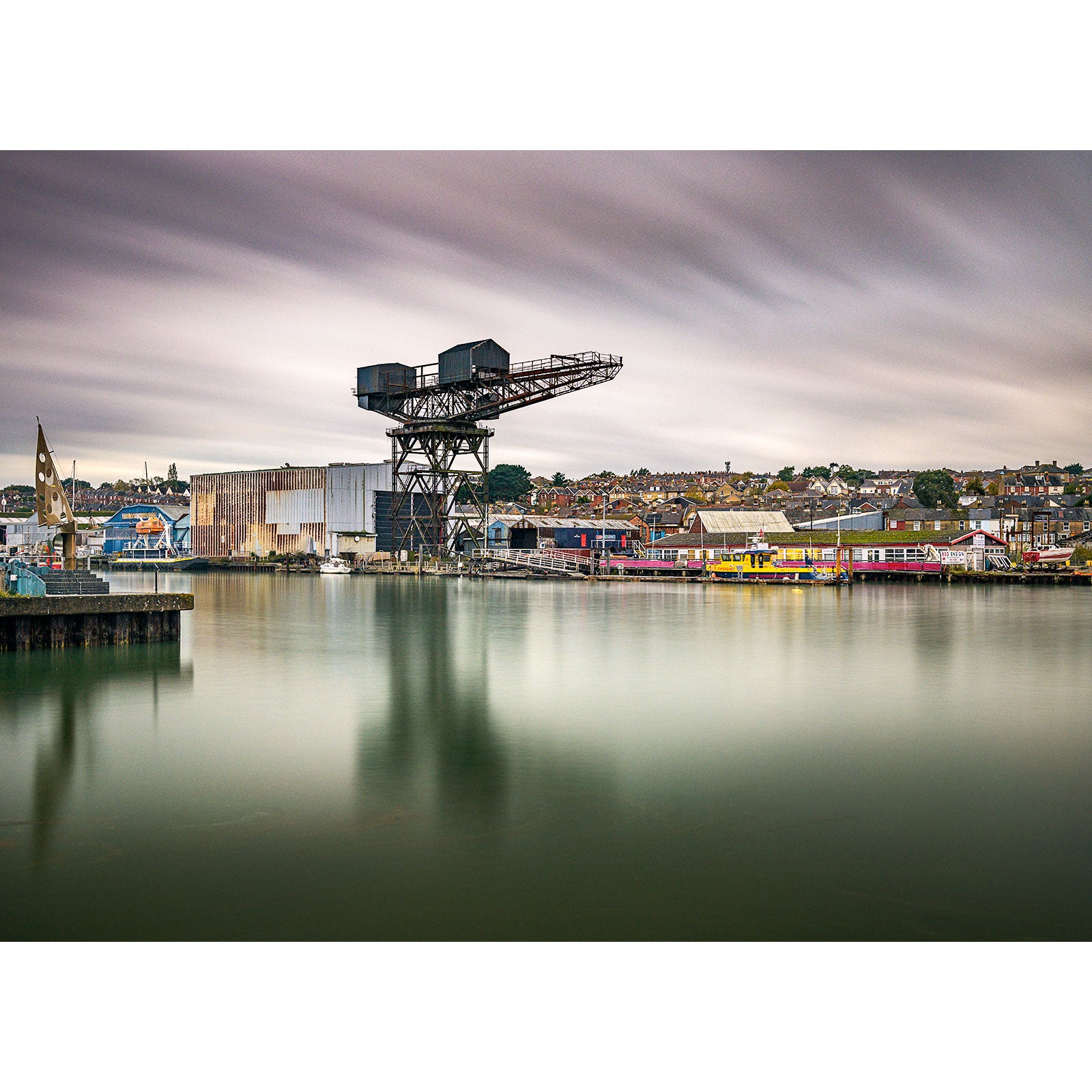 Industrial waterfront scene on the Isle, with a Hammerhead Crane overlooking a calm river, under a cloudy sky. (Brand: Available Light Photography)