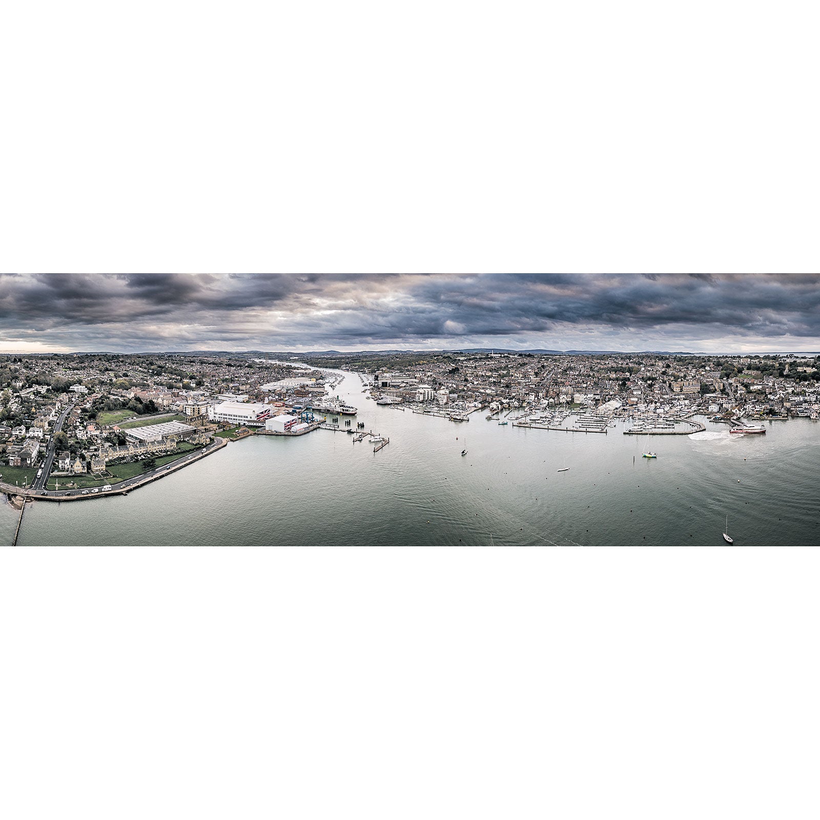 A panoramic aerial view of Cowes and East Cowes on the Isle of Wight with a harbor, boats, and surrounding residential areas under a cloudy sky by Available Light Photography.