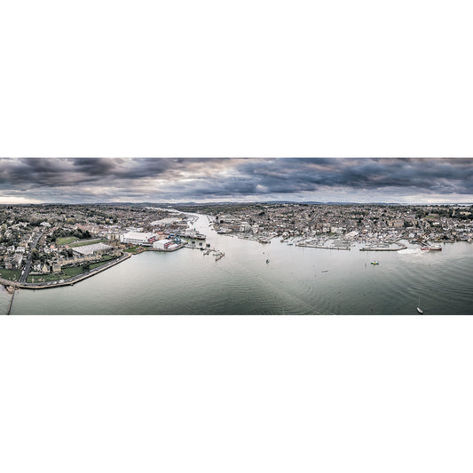 A panoramic aerial view of Cowes and East Cowes on the Isle of Wight with a harbor, boats, and surrounding residential areas under a cloudy sky by Available Light Photography.