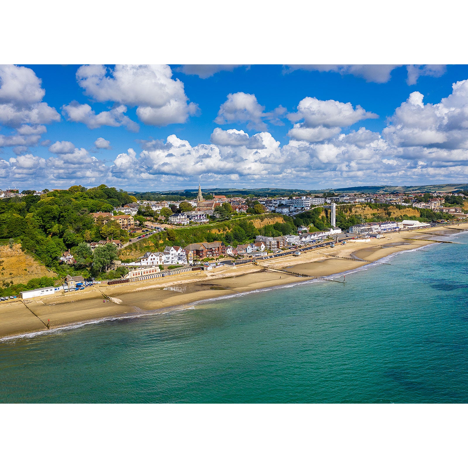 Aerial view of a coastal town with a beachfront, buildings, and greenery under a partly cloudy sky taken with the Shanklin by Available Light Photography.