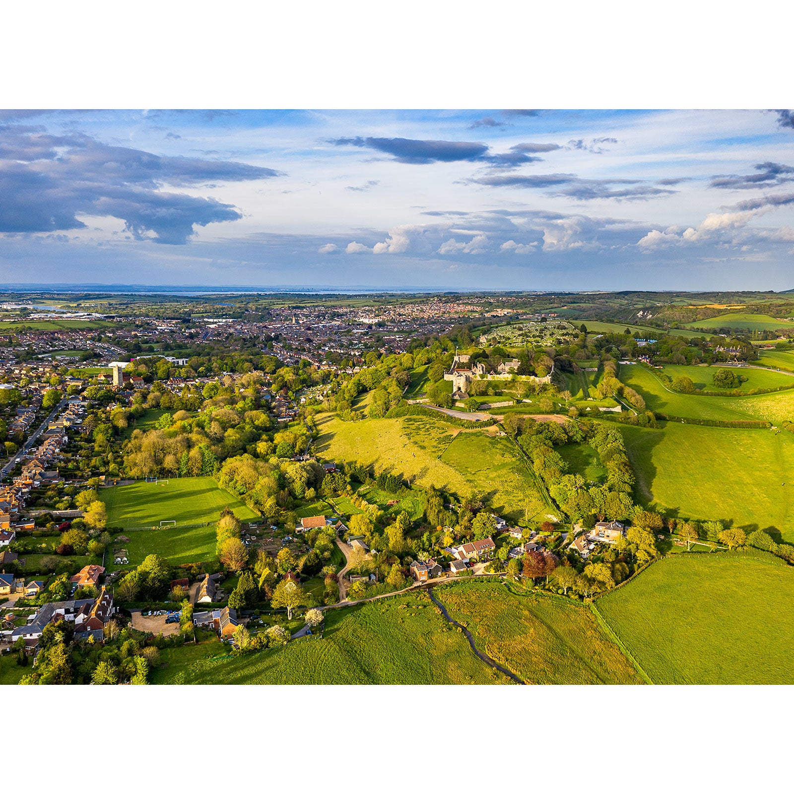 Aerial view of a lush green landscape with Carisbrooke town amidst rolling hills under a cloudy sky by Available Light Photography.