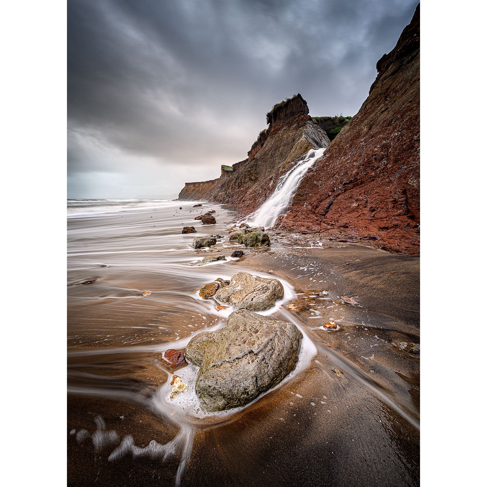 A rugged coastline with steep cliffs and rock formations under a cloudy sky, with waves washing over a sandy beach on the Isle of Wight photographed by Available Light Photography featuring Dinosaur Footprints, Brook.