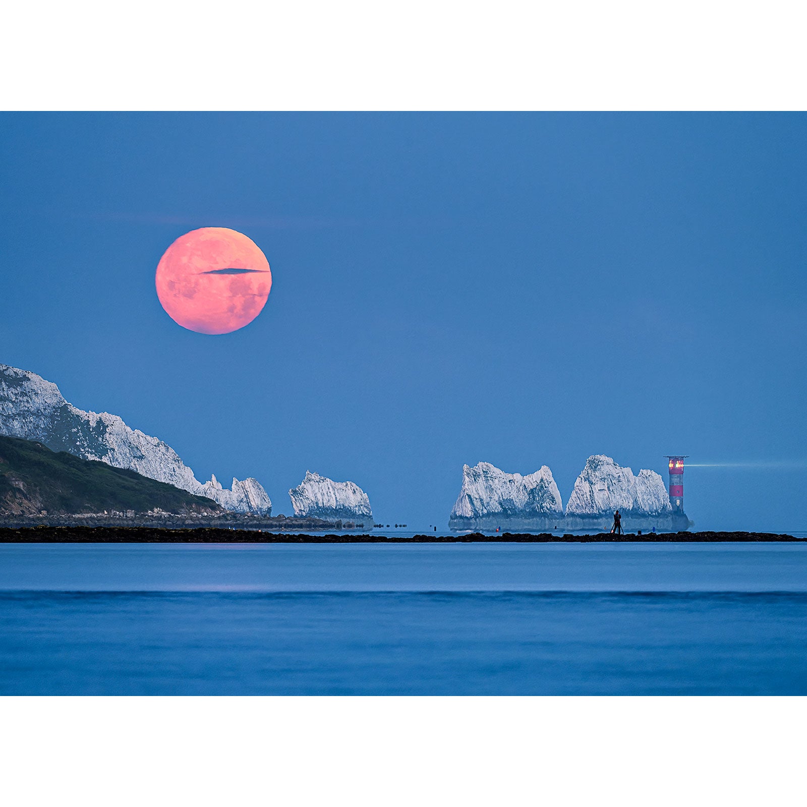 Moonset over The Needles rising over a tranquil sea beside a rocky coast with a lighthouse on the Isle of Wight by Available Light Photography.