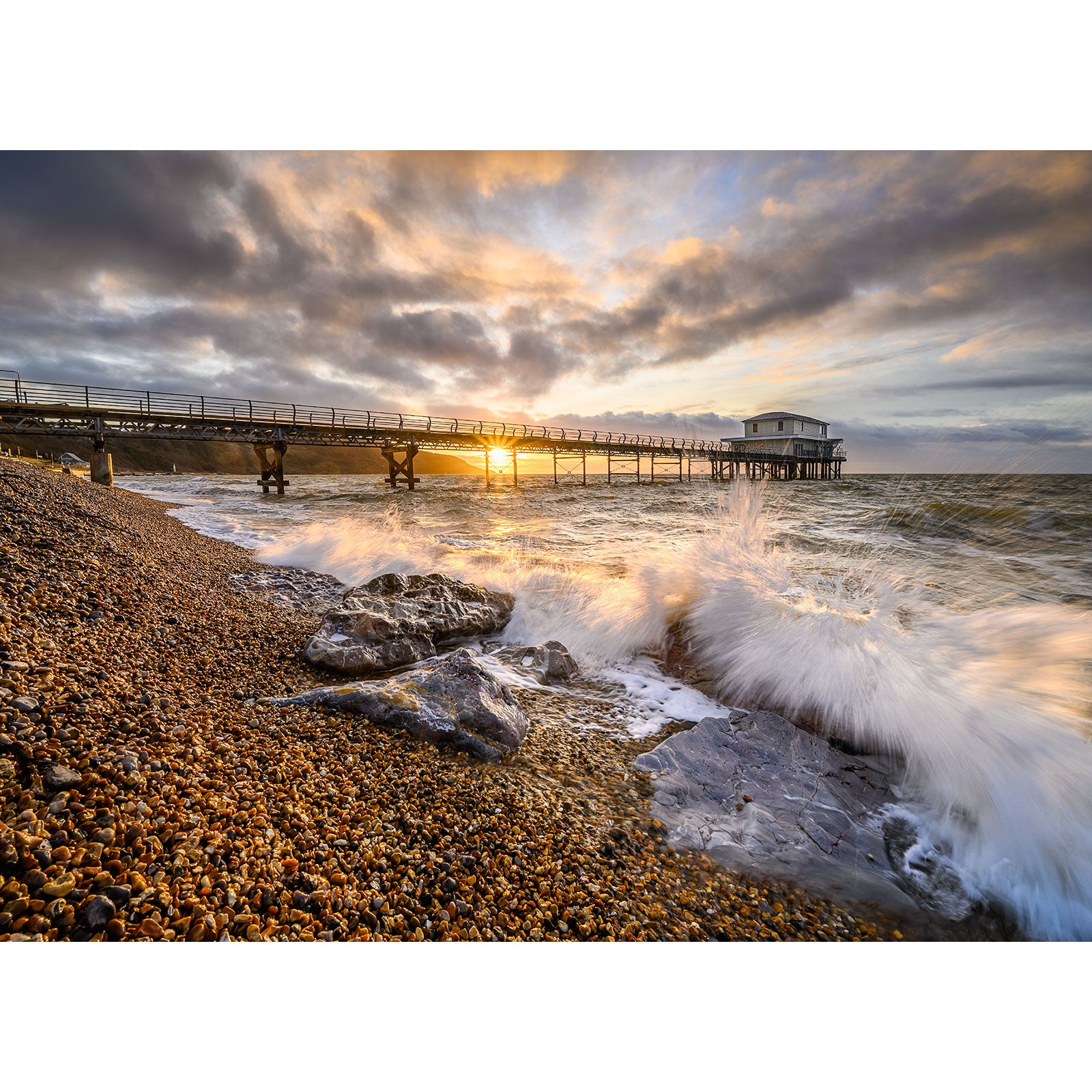 A serene sunset at a Totland beach with waves crashing against rocks on the Isle of Wight, and a pier extending into the sea by Available Light Photography.